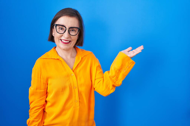 Mujer hispana de mediana edad con gafas de pie sobre fondo azul sonriendo alegre presentando y señalando con la palma de la mano mirando a la cámara.  - Foto, Imagen