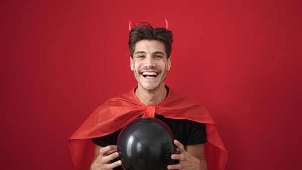 jonge latino man dragen duivel kostuum houden ballon over geïsoleerde rode achtergrond - Foto, afbeelding