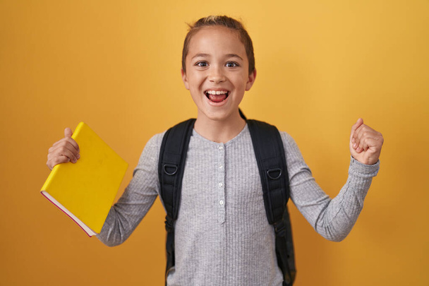 Little caucasian boy wearing student backpack and holding book screaming proud, celebrating victory and success very excited with raised arms  - Fotoğraf, Görsel
