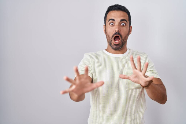 Hispanic man with beard standing over isolated background afraid and terrified with fear expression stop gesture with hands, shouting in shock. panic concept.  - Photo, Image