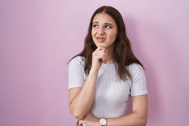 Junges hispanisches Mädchen, das über rosa Hintergrund steht und besorgt über eine Frage nachdenkt, besorgt und nervös mit der Hand am Kinn  - Foto, Bild