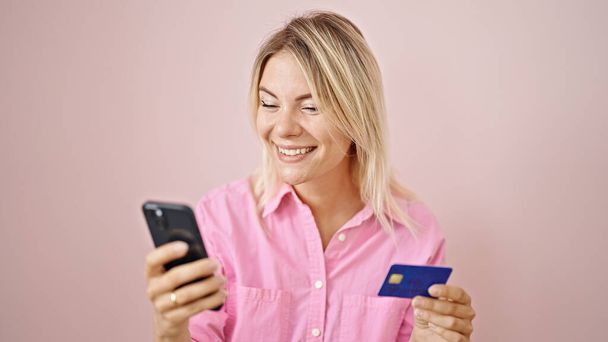 Young blonde woman shopping online with smartphone and credit card over isolated pink background - Foto, Imagem