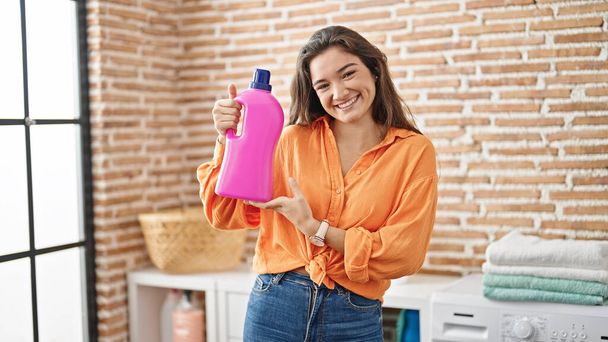 Jonge mooie Spaanse vrouw met fles wasmiddel glimlachend in de wasruimte - Foto, afbeelding