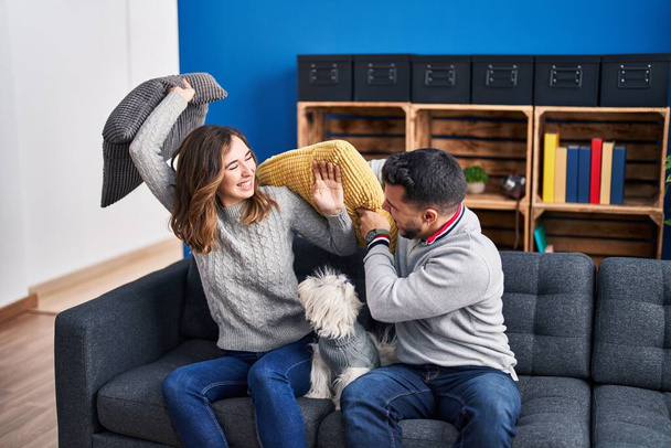 Man and woman fighting with cushion sitting on sofa with dog at home - Fotó, kép