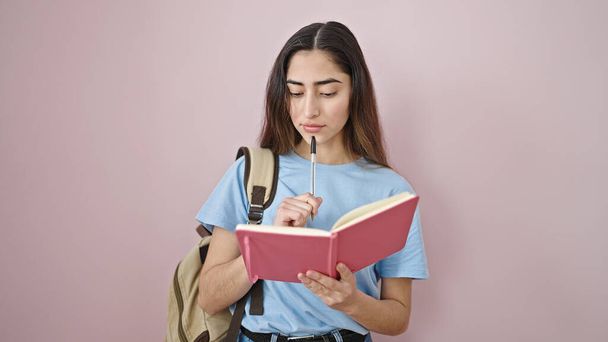 Νεαρή όμορφη ισπανόφωνη μαθήτρια που γράφει στο σημειωματάριο πάνω από απομονωμένο ροζ φόντο - Φωτογραφία, εικόνα