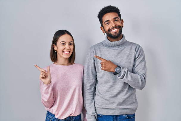 Mladý hispánský pár stojí spolu veselý s úsměvem na tváři ukazuje rukou a prstem nahoru na stranu se šťastným a přirozeným výrazem  - Fotografie, Obrázek
