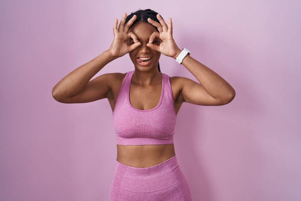 ピンクの背景にスポーツウェアを着たアフリカ系アメリカ人の女性は、双眼鏡が舌を突き出しているようなokジェスチャーをし、目は指を通して見ている。クレイジーな表現.  - 写真・画像