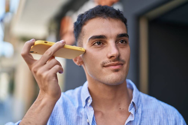 Jonge Spaanse man glimlacht vol vertrouwen luisterend audioboodschap door de smartphone op straat - Foto, afbeelding