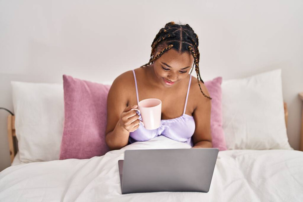 Αφροαμερικανή γυναίκα που χρησιμοποιεί φορητό υπολογιστή πίνοντας καφέ στο υπνοδωμάτιο - Φωτογραφία, εικόνα