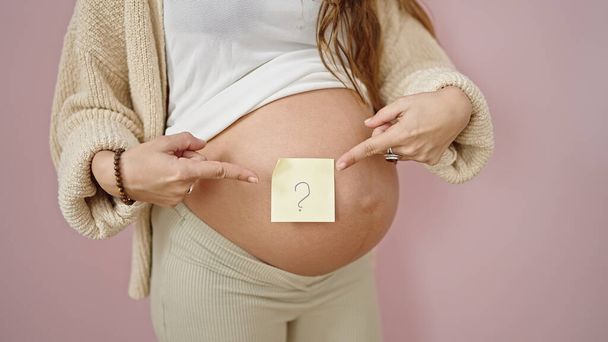 Νεαρή έγκυος γυναίκα δείχνει με το δάχτυλο στο ερωτηματικό σημείωμα χαρτί υπενθύμισης στην κοιλιά πάνω από απομονωμένο ροζ φόντο - Φωτογραφία, εικόνα