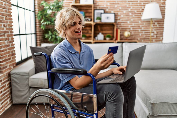 Νεαρός ξανθός άντρας που χρησιμοποιεί φορητό υπολογιστή και πιστωτική κάρτα κάθεται σε αναπηρικό καροτσάκι στο σπίτι - Φωτογραφία, εικόνα