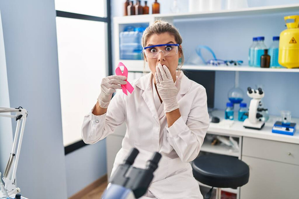 Eine junge Frau, die im wissenschaftlichen Labor arbeitet, hält ein rosafarbenes Band in der Hand, das den Mund mit der Hand bedeckt, schockiert und aus Angst vor Irrtümern. Überraschte Miene  - Foto, Bild
