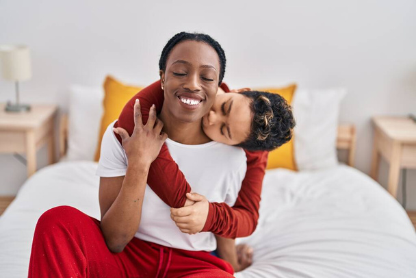 Afrikanisch-amerikanische Mutter und Sohn umarmen einander und küssen sich im Schlafzimmer auf dem Bett sitzend - Foto, Bild