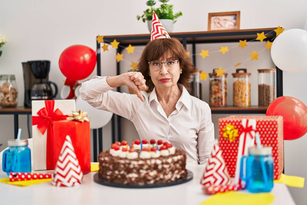 Жінка середнього віку, яка святкує день народження, тримає великий шоколадний торт з сердитим обличчям, негативний знак, що показує нелюбов до великих пальців вниз, концепція відторгнення
  - Фото, зображення