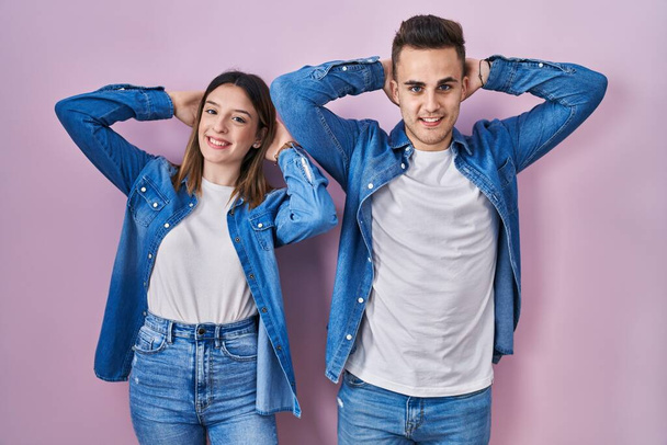 Fiatal spanyol pár áll a rózsaszín háttér pihentető és nyújtózkodó, karok és kezek a fej és a nyak mögött mosolyogva boldog  - Fotó, kép