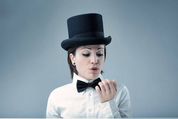 Femme au chapeau haut de forme, air de supériorité, soufflant sur ses ongles, n'exige rien de moins que ses normes. Chemise blanche, noeud papillon noir, fond neutre - Photo, image