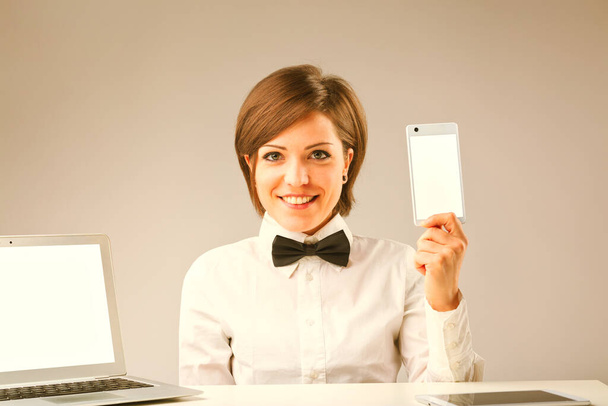 Vintage-style εικόνα μιας γυναίκας που παρουσιάζει το διαδίκτυο και ψηφιακές συσκευές με άφθονο COPYSPACE. Φορώντας ένα λευκό πουκάμισο και παπιγιόν, επιδεικνύει ένα smartphone και έχει ένα φορητό υπολογιστή και ένα ψηφιακό tablet σε αυτόν - Φωτογραφία, εικόνα