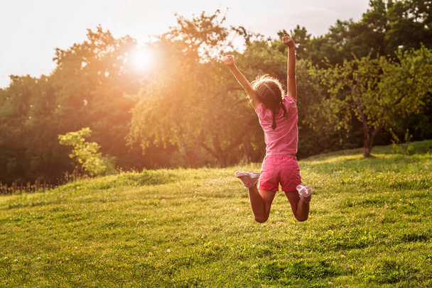 Viel Spaß im Sommerurlaub vor dem Hintergrund der Natur. Kleines Mädchen hüpft und genießt das Leben auf grünem Gras mit erhobenen Händen im Sonnenuntergang. - Foto, Bild
