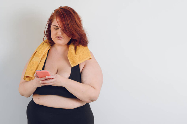 young plus size argentinian ethnicity hispanic latina woman with red hair and black sportswear standing using phone over white background, technology concept, copy space. - Fotó, kép