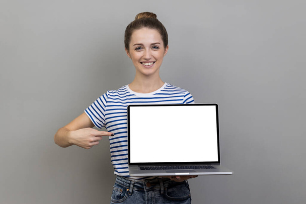 Portrait of woman wearing striped T-shirt pointing finger at laptop with white blank display, showing empty screen, bragging with internet advertisement. Indoor studio shot isolated on gray background - Foto, Bild
