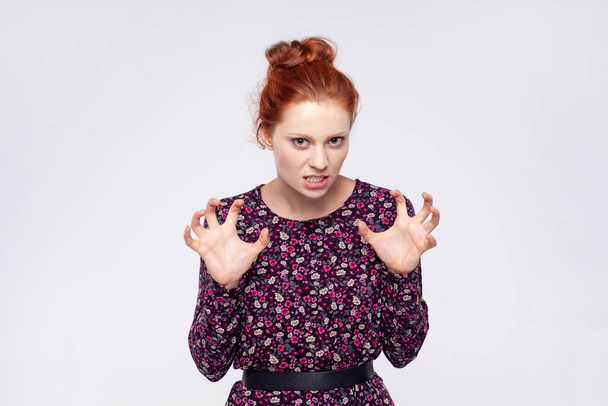Portrait of angry aggressive young adult ginger woman wearing dress standing with raised arms and clenching teeth, expressing hate and anger. Indoor studio shot isolated on gray background. - Photo, Image