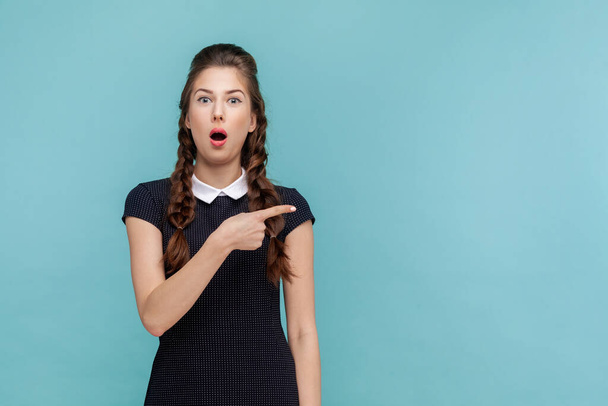 Portrait of shocked surprised woman points away on copy space, suggests follow this direction or click on link, wearing black dress. woman Indoor studio shot isolated on blue background. - 写真・画像