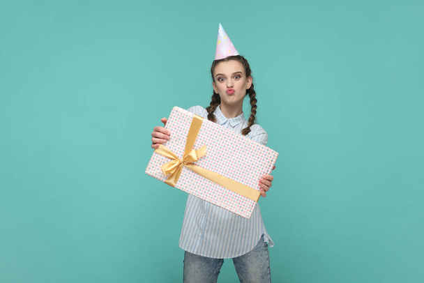 Portrait of funny positive teenager girl with braids wearing striped shirt and party cone, holding gift in wrapped box, looks at camera with pout lips. Indoor studio shot isolated on green background. - Fotografie, Obrázek