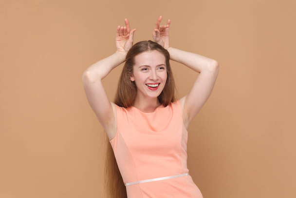 Portrait of funny smiling joyful woman with long hair standing with raised arms, showing horns, looking at camera, wearing elegant dress. Indoor studio shot isolated on brown background. - Photo, image