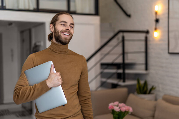 Smiling attractive man with beard wearing casual clothing standing at home with folded laptop. - Photo, Image