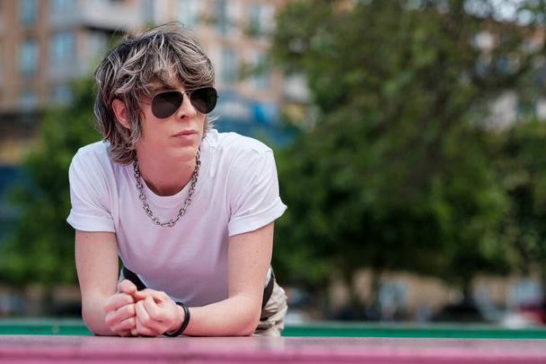 Portrait of androgynous female leaning on a pink table outdoors. She is in a park wearing sunglasses and white t-shirt. Non -binary, lgbtq, diversity and freedom concept. - Photo, image