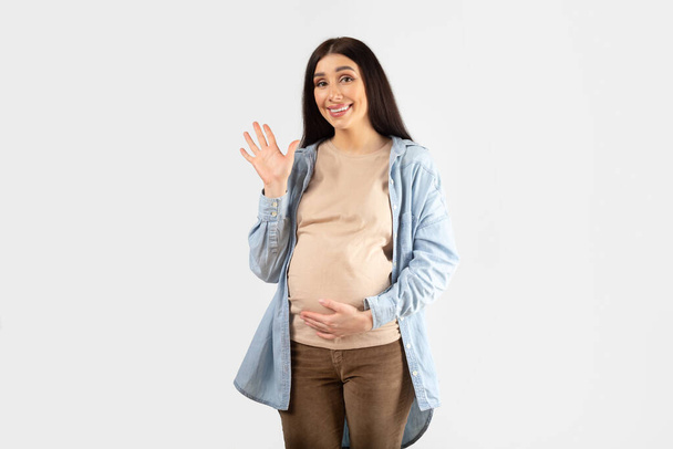 Positive young expectant woman touching belly and waving at camera saying hello and smiling, posing on white studio background. Friendly welcome gesture - Photo, Image