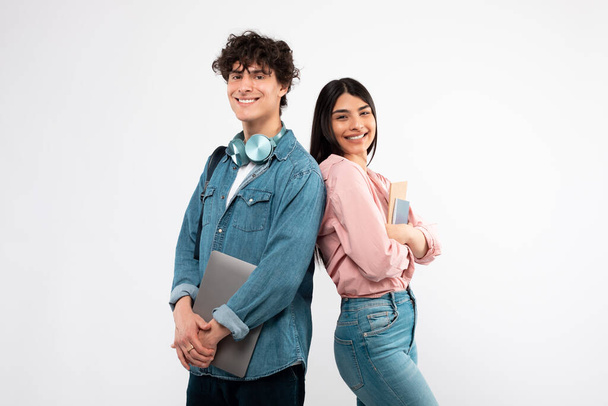 Moderne Studenten. Glückliches junges Paar mit Laptop und Arbeitsbüchern posiert Rücken an Rücken auf weißem Studiohintergrund und lächelt in die Kamera. Technologie und Bildung, tolles E-Learning-Angebotskonzept - Foto, Bild