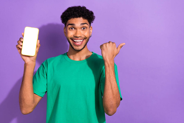 Portrait of amazed handsome guy hold show empty space smart phone screen direct finger offer isolated on purple color background. - 写真・画像