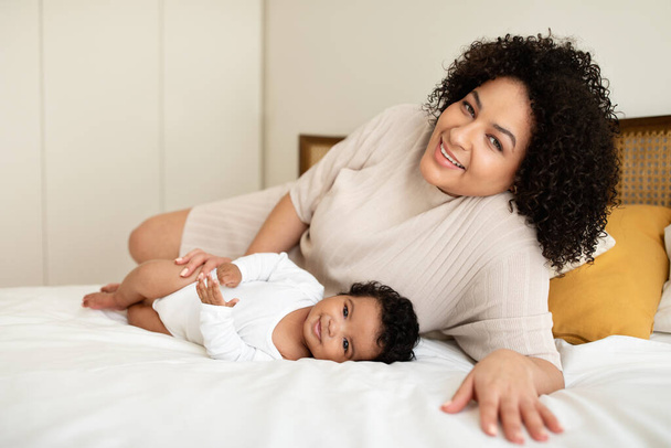 Child care vacation at home. Cheerful black millennial mother plays with little baby on white bed, enjoys love, rest and free time in bedroom interior. Parenthood and family, ad and offer - Photo, Image