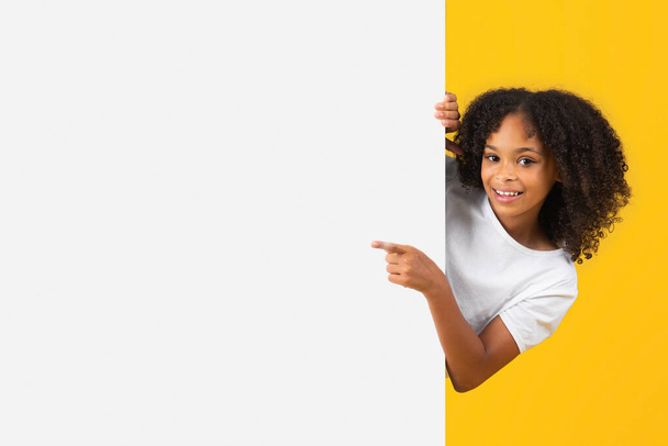 Cheerful adolescent black curly girl in white t-shirt point finger on big banner with empty space, recommends ad and offer, isolated yellow studio background. Knowledge, study, pupil emotions, sale - Foto, Imagen