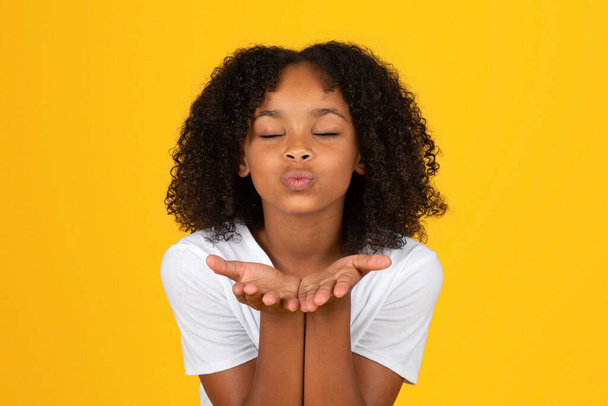 Happy confident adolescent curly girl in white t-shirt with closed eyes makes blowing kiss in camera, isolated on yellow studio background. Love, romance, student, knowledge and education in school - Φωτογραφία, εικόνα