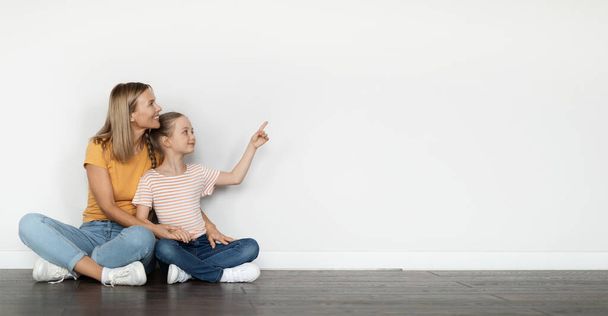 Spójrz na to. Cute Little Girl wskazując poza podczas relaksu z mamą na podłodze, szczęśliwy matka i córka Wyświetlanie skopiować miejsce na reklamę siedząc w pobliżu białej ściany w domu, Panorama - Zdjęcie, obraz