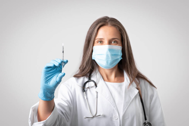 Senior woman therapist in white coat and protective mask showing syringe with vaccine, isolated on gray background, selective focus. Lékař provádějící očkování, očkování pro zdravotní péči - Fotografie, Obrázek