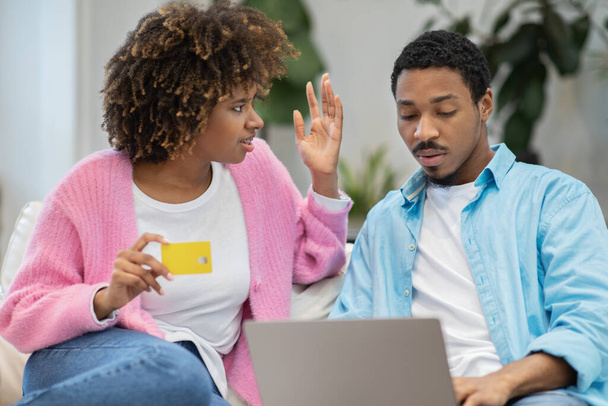 Financial disagreement in couple, marriage, relationships. Unhappy black couple sitting on couch at home, using laptop and bank card, spouses have quarrel while shopping or banking online - Photo, image