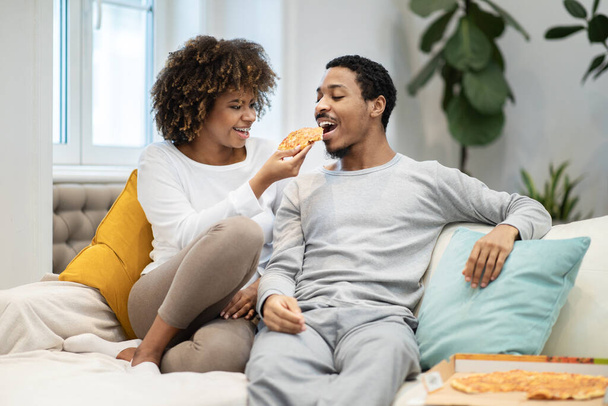 Щасливі красиві тисячолітні чорні коханці сидять на дивані, їдять піцу, дружина годує чоловіка, люблять афроамериканця і жінку, проводять вихідні разом вдома, копіюють простір
 - Фото, зображення