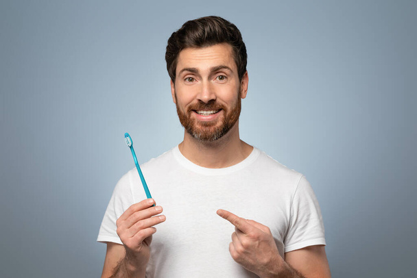 Bright smile ritual. Cheerful middle aged man pointing at toothbrush and smiling at camera on grey studio background. Male taking care of his teeth, doing morning routine - Photo, image
