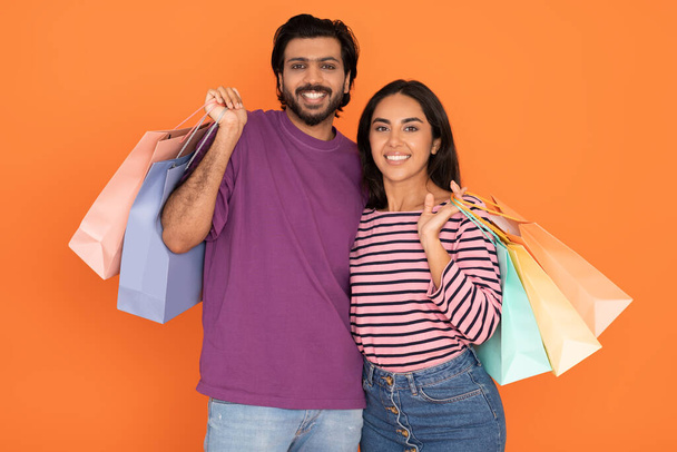 Retail, black friday, season sale. Happy smiling young eastern couple in casual outfits embracing, posing with colorful shopping bags isolated on orange studio background - Photo, Image