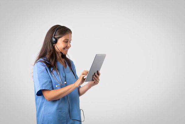 Telemedicine concept. Cheerful senior woman doctor with tablet and headset making online consultation, physician talking to patient via video call on device, grey background, free space - Photo, Image