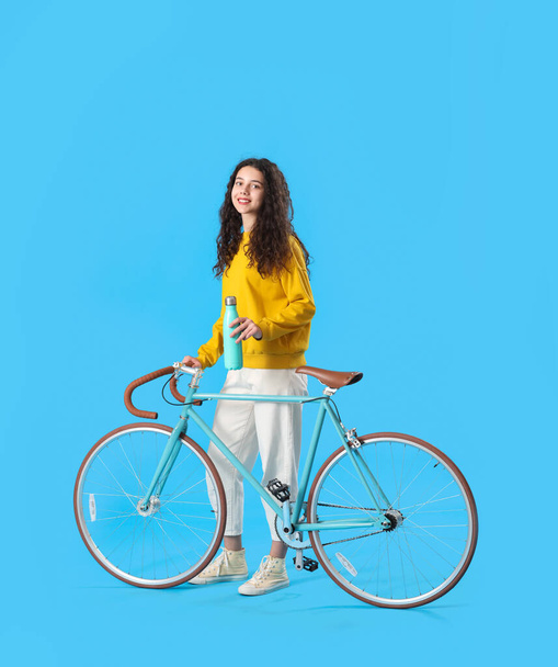 Teenage girl with bottle of water and bicycle on blue background - Фото, зображення