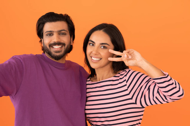 Studio portrait of cheerful attractive millennial indian couple wearing casual outfit embracing, taking selfie together on orange studio background, showing peace gesture, smiling at camera - Zdjęcie, obraz