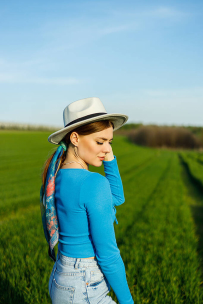 Una mujer linda en jeans y un sombrero se encuentra en un campo verde. Una mujer sonriente con un top azul y jeans camina en la hierba verde. - Foto, Imagen