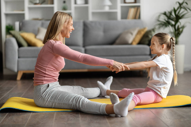 Urheilutoiminta. Onnellinen perhe äiti ja pieni tytär harjoittelemassa yhdessä kotona, iloinen nainen ja hänen söpö naispuolinen lapsi tekemässä kuntoilua olohuoneessa sisustus, kädet kädessä ja hymyily - Valokuva, kuva