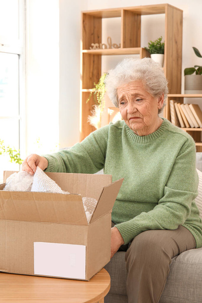 Upset senior woman opening parcel at home - Foto, afbeelding