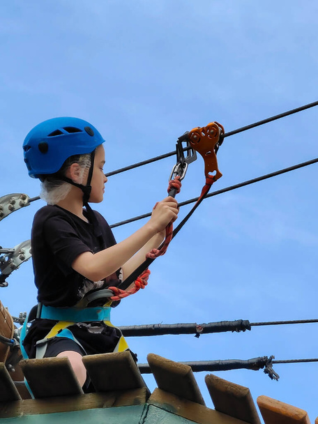 Un adolescent portant un casque bleu de sécurité sportive et un équipement de sécurité attache un mousqueton à un téléphérique dans un parc d'attractions à cordes - Photo, image