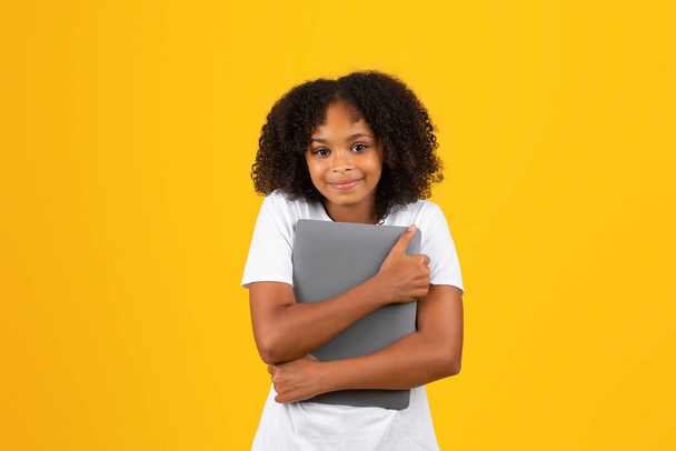 Happy adolescent curly black girl in white t-shirt hugs tablet, isolated on yellow studio background. Device for blog and website for education, study, love to social networks and chat - Foto, Bild